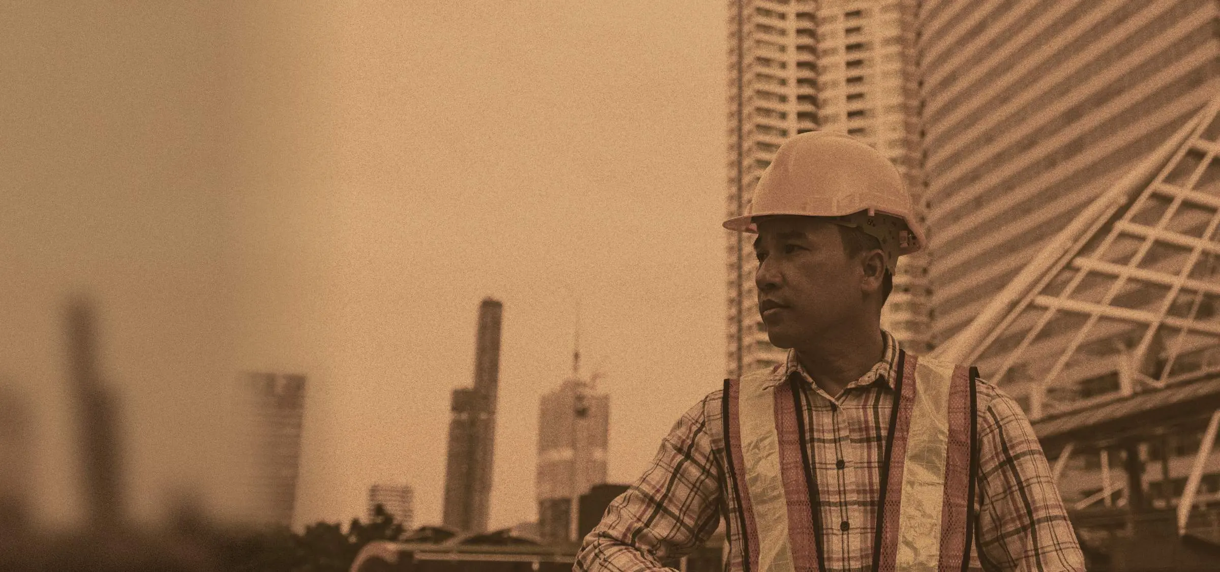 Pria berhelm proyek dan rompi keselamatan menoleh ke samping, dengan gedung tinggi di belakangnya