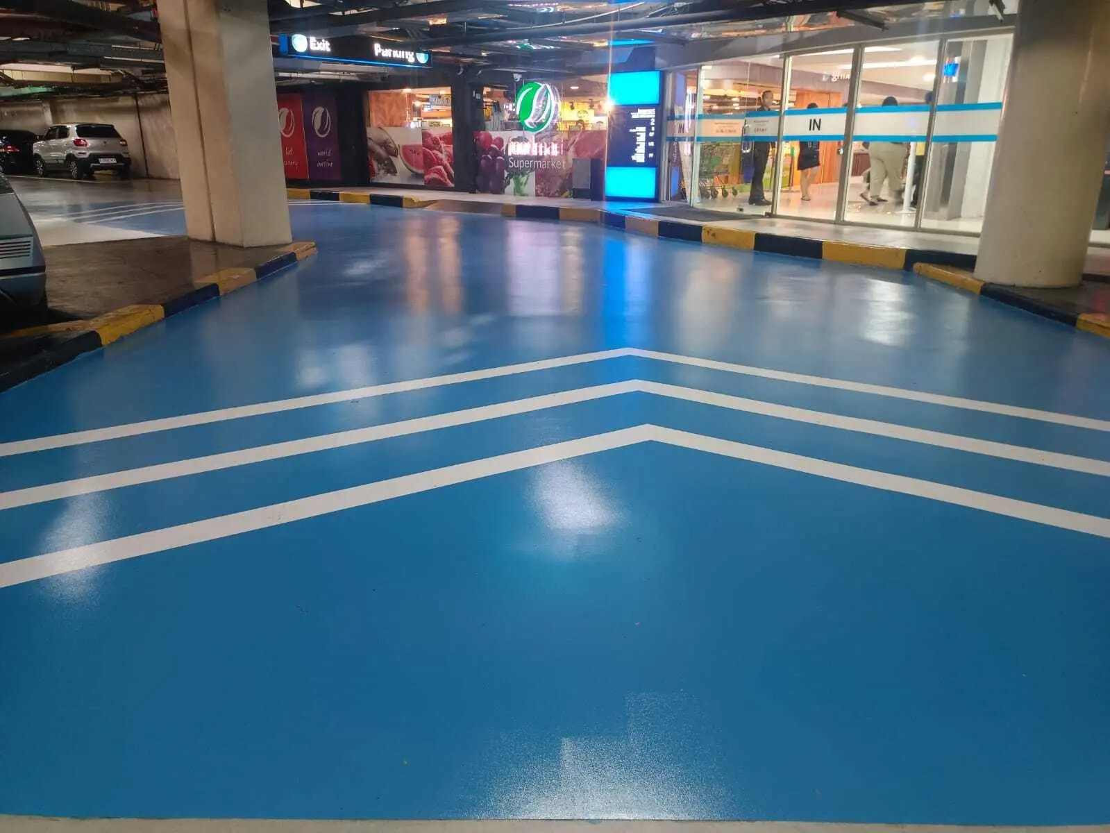 Jalur masuk parkir mal dengan epoxy biru dan marka chevron putih di depan pintu supermarket
